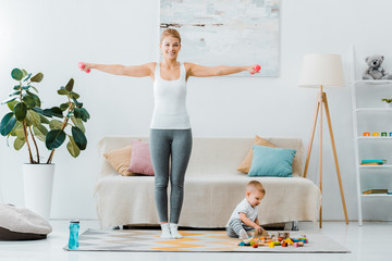 smiling woman doing exercise with dumbbells and looking at camera and toddler boy playing with multicolored cubes on carpet in living room