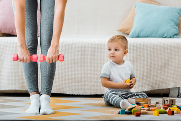 Naklejka premium cropped view of woman doing fitness exercise with dumbbells and cute toddler boy sitting on carpet near multicolored cubes