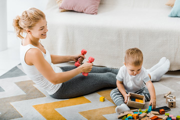 attractive woman exercising with dumbbells and looking at cute toddler boy holding wooden box with multicolored cubes near couch