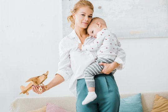 attractive woman looking at camera and holding wooden plane model and toddler boy in living room