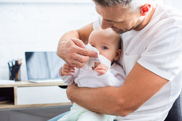 father wiping runny nose of adorable baby daughter with napkin at home