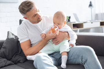 handsome father sitting on couch and feeding baby daughter from bottle at home