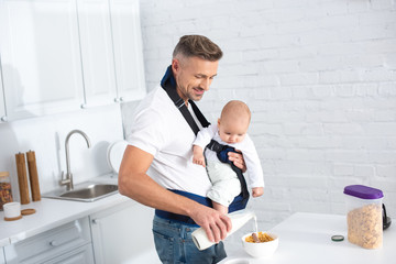 father holding infant daughter in baby carrier and pouring milk in bowl with cornflakes