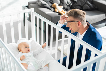 happy father in formal wear showing tongue to infant daughter in baby crib - obrazy, fototapety, plakaty