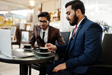 Two indian business man in suits sitting at office on cafe, looking at laptop and drinking coffee.