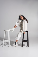 attractive african american girl in stylish white clothes and hat sitting on black and white chairs on white