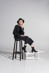 beautiful caucasian blonde woman in black clothes and hat sitting on chair and looking at camera on white