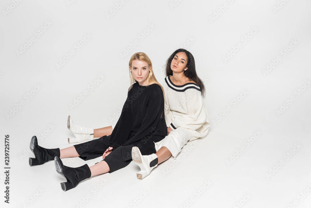 Wall mural beautiful multicultural women in black and white clothes sitting and looking at camera on white