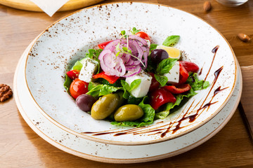 Greek salad with feta cheese, olives and herbs, served in a white plate on a table in a restaurant.