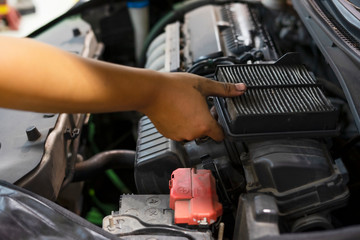An auto mechanic wearing protective work gloves holds a dirty, clogged air filter over a car engine...