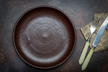 Empty brown plate (ceramic) on a dark background with a knife and fork. Copy space.