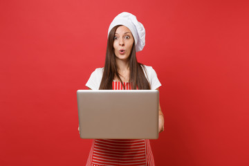 Housewife female chef cook or baker in striped apron, white t-shirt, toque chefs hat isolated on red wall background. Fun housekeeper woman looking for recipe in laptop pc. Mock up copy space concept.