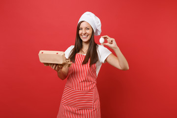 Housewife female chef cook or baker in striped apron t-shirt toque chefs hat isolated on red wall background. Woman hold brown craft cardboard container for chicken eggs. Mock up copy space concept.