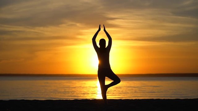 Silhouette girl doing tree yoga pose at sunset in nature outdoors. Sporty Yoga woman with open raised arms practicing yoga in sea water beach. Female yoga working out training through shining sun rays