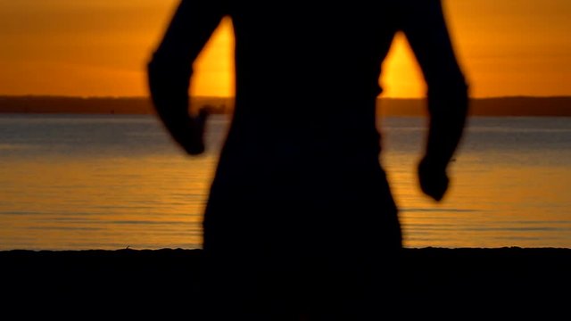 Silhouette of sporty woman running in sea ocean beach at sunset. Female jogger jogging in nature. Athletic girl running outdoor. Healthy fitness wellness sport concept. Sun rays slow-motion