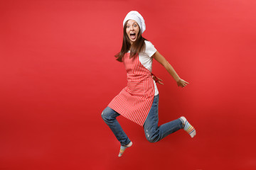 Housewife female chef cook or baker in striped apron white t-shirt, toque chefs hat isolated on red wall background. Full length portrait housekeeper woman jumping high up. Mock up copy space concept.