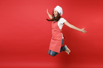 Housewife female chef cook or baker in striped apron white t-shirt, toque chefs hat isolated on red wall background. Full length portrait housekeeper woman jumping high up. Mock up copy space concept.
