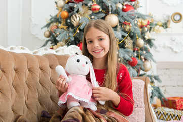 Best Christmas toy. Little girl with cute bunny at Christmas tree. Small girl hold rabbit toy. Little child play with soft toy. Small child happy smiling with present. The most anticipated toy