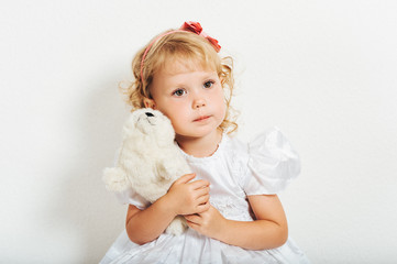 Studio shot of adorable 3 year old toddler girl wearing white occasion dress, holding soft plush toy