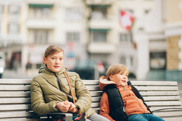 Outdoor portrait of two kids wearing warm jackets, sitting on the bench, spring fashion for children