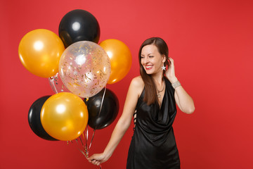 Fototapeta premium Pretty young woman in little black dress celebrating holding air balloons isolated on red background. St. Valentine's, International Women's Day, Happy New Year, birthday mockup holiday party concept.