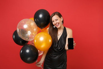 Smiling young woman in little black dress celebrating holding mobile phone with blank black empty screen, air balloons isolated on red background. Happy New Year birthday mockup holiday party concept.