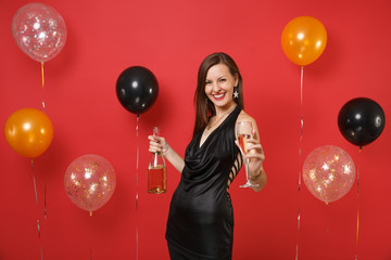 Laughing young girl in black dress celebrating holding glass, bottle of champagne on bright red background air balloons. International Women's Day Happy New Year birthday mockup holiday party concept.