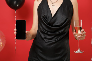 Cropped image of girl in black dress holding glass of champagne, mobile phone with blank black empty screen on bright red background air balloons. Happy New Year birthday mockup holiday party concept.