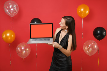 Pretty young woman in black dress looking on laptop pc computer with blank black empty screen in hands on bright red background air balloons. Happy New Year, birthday mockup holiday party concept.