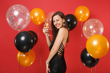 Side view of smiling pretty girl in black dress celebrating, holding glass of champagne on bright red background air balloons. Valentine's Day, Happy New Year, birthday mockup holiday party concept.