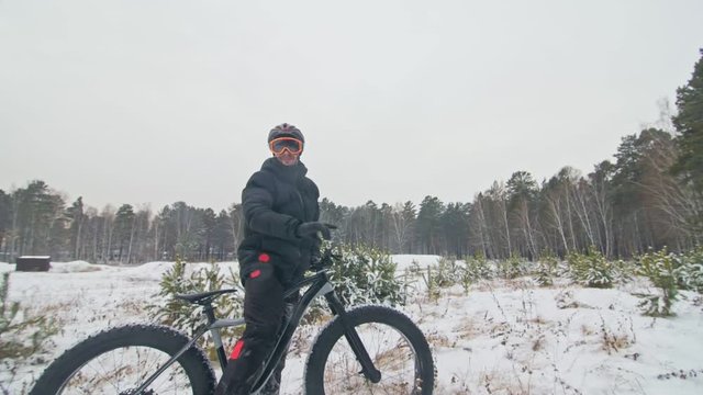 Professional extreme sportsman biker stand a fat bike in outdoors. Cyclist recline in the winter snow forest. Man walk with mountain bicycle with big tire in helmet and glasses. Slow motion in 180fps.