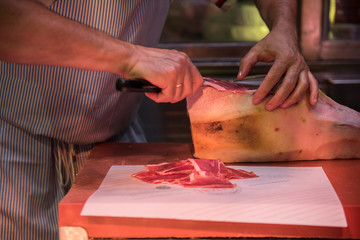 A butcher is seen cutting jamon meat with a butcher knife. The jamon looks fresh and is ready to be sold after being cut. He is seen wearing a stripe Apron.