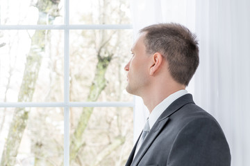 Closeup profile, side of young man, male businessman in business gray, grey suit, tie, bridegroom, standing in home, house, apartment room, white curtains, looking through window on view, thinking