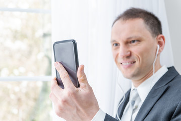 Closeup of young, happy man, male businessman in business suit, tie, standing by window in home, house room, white curtains, with earbuds headphones, holding phone, talking, video conference