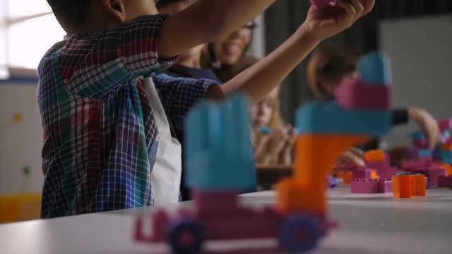 Cheerful mixed race preschool boy playing with toys made of colorful construction blocks in kindergarten over attractive female teacher and diverse multicultural children busy in background.