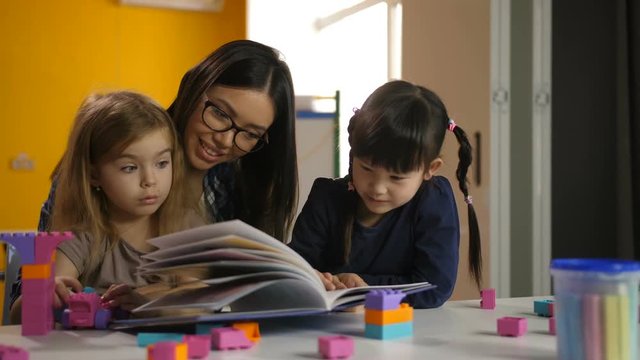Portrait of gorgeous preschool female teacher in eyeglasses with adorable multinational girls looking through picture book in kindergarten. Positive teacher reading a book to little pupils at class.