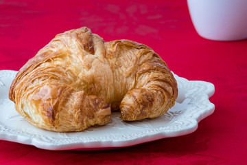Fresh croissant on a white stoneware plate on red tablecloth, with white coffee cup