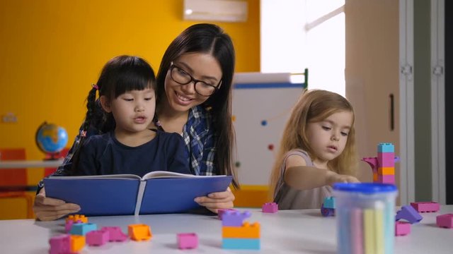 Cheerful preschool asian girl with mixed race teacher learning and reading children's picture book in kindergarten class while cute caucasian little girl playing with colorful construction toy blocks.