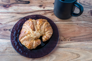 Fresh croissant on a purple glass plate on a wood cutting board background with black coffee cup, top view