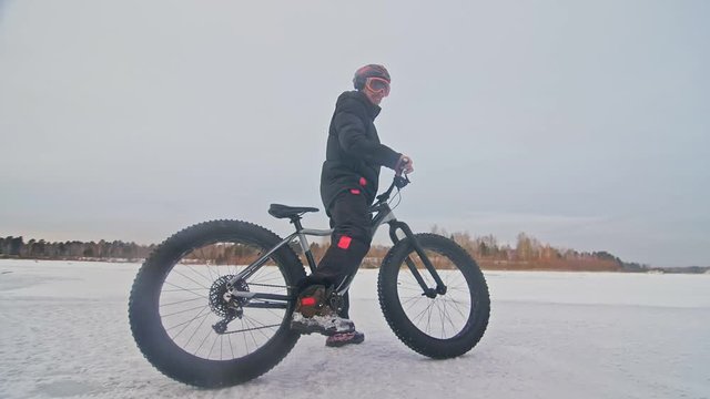 Professional extreme sportsman biker stand a fat bike in outdoors. Cyclist recline in the winter snow forest. Man walk with mountain bicycle with big tire in helmet and glasses. Slow motion in 180fps.