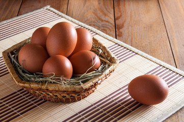 Group of large brown chicken eggs lies in a wicker basket and one lies next to the basket, on a wooden table.