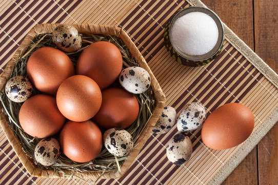 Many Eggs. Large Brown Chicken Eggs And Small Motley Quail Eggs On Straw In Wicker Basket And Salt In Salt Shaker On Wooden Table. View From Above.