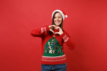 Joyful pretty young Santa girl in knitted sweater, Christmas hat showing shape heart with hands isolated on red background. Happy New Year 2019 celebration holiday party concept. Mock up copy space.