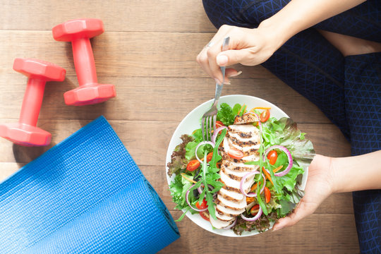 Top view healthy girl eating chicken salad. Dumbbells and yoga mat on the floor