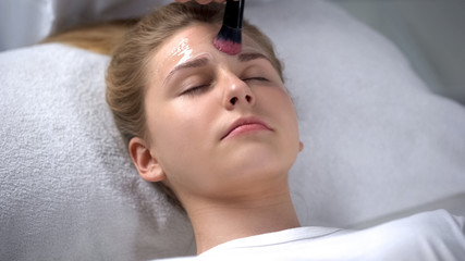 Obraz na płótnie Canvas Worker of beauty salon putting moisturizing mask on girl face, femininity