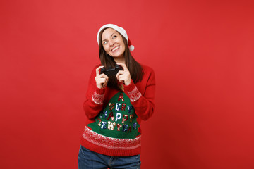 Dreamy young Santa girl in sweater Christmas hat looking up playing video game with joystick isolated on red wall background. Happy New Year 2019 celebration holiday party concept. Mock up copy space.