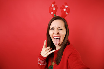 Close up selfie shot of Santa girl in fun decorative deer horns showing horns gesture depicting heavy metal rock sign isolated on red background. Happy New Year 2019 celebration holiday party concept.