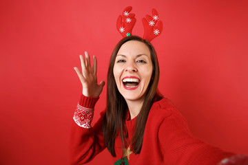 Close up selfie shot of joyful young Santa girl in fun decorative deer horns rising hand isolated on bright red background. Happy New Year 2019 celebration holiday party concept. Mock up copy space.