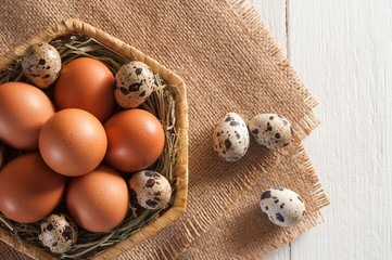 Several large brown chicken eggs and small motley quail eggs on burlap in wicker basket. Near three quail eggs. White wooden background.