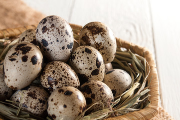 Many quail eggs on straw in wicker basket. White wooden background. Healthy food.